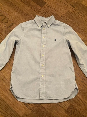 Ljusblå skjorta från Polo Ralph Lauren - Snygg ljusblå skjorta från Polo Ralph Lauren med klassisk button-down krage och broderad logga på bröstet. Skjortan har långa ärmar och knappar framtill. Perfekt för dig som gillar stilrena och tidlösa plagg.