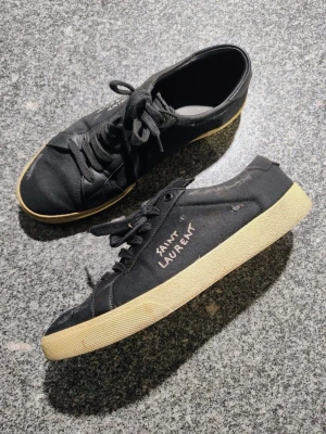 Svarta sneakers från Saint Laurent - Svarta sneakers från Saint Laurent med beige platt gummisula och snörning. Ovandelen är i canvas med detaljer i skinn på hälen. Saint Laurent-logga syns på sidan och bak på hälen. Rund tå och klassisk låg profil, perfekt för en clean streetstil.
