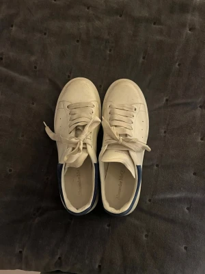 Alexander McQueen vita sneakers - Säljer ett par vita sneakers från Alexander McQueen med blå mockadetalj vid hälen. Skorna har snörning, rund tå och platt sula med unikt mönster undertill. Tillverkade i skinn och mocka, klassisk och chunky design som är riktigt snygg till jeans eller kjol.