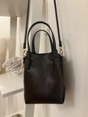 Mörkbrun axelväska - Mörkbrun axelväska med guldfärgade metalldetaljer. Väskan har både korta handtag och en lång justerbar axelrem som även går att ta av 👜