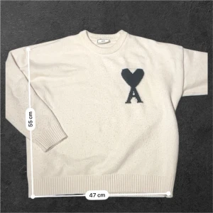 Vit stickad tröja från Ami - Säljer en vit stickad tröja från Ami med ett mörkblått hjärta och bokstaven A broderat på bröstet. Tröjan har rund hals, ribbade muddar och långärmad passform. Perfekt för dig som gillar stilrena och trendiga plagg.