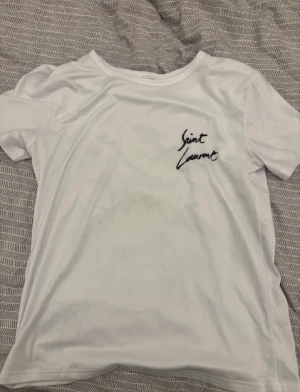 Vit Saint Laurent t-shirt med tryck - Säljer en vit t-shirt från Saint Laurent med svart handskrivet tryck både fram och bak. T-shirten har rund hals och korta ärmar, tillverkad i mjuk bomull som känns skön mot huden. Perfekt för dig som gillar stilrena designer med en touch av lyx.