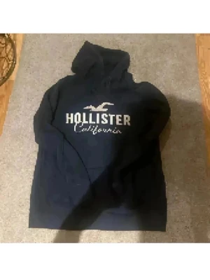 Hollister Hoodie💕 - Svart hoodie från Hollister med stor vit logga och texten 'California' på bröstet. Tröjan har huva med snörning och är långärmad. Perfekt för dig som gillar en enkel och klassisk streetstil. Materialet känns mjukt och bekvämt, troligen bomull eller polyester. Strl L på den men passar super oversize på xs-m