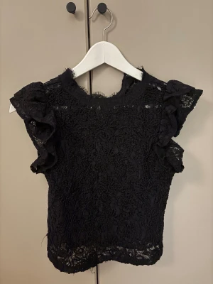 Svart spetsblus från Zara - Säljer en svart blus från Zara i storlek S. Blusen är gjord i spets med blommigt mönster och har volangdetaljer på axlarna. Den är ärmlös och har en rund hals med lite rå kant. Perfekt för dig som gillar detaljer och en elegant look.
