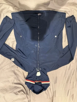 Mörkblå vindjacka från Moncler - Snygg mörkblå vindjacka från Moncler med huva och dragkedja framtill. Jackan har Moncler-logga på ärmen och coola detaljer som rödvit rand på huvan. Tillverkad i polyester och har praktiska fickor med tryckknappar. Perfekt för dig som gillar stil och funktion.