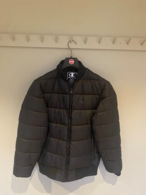 Svart pufferjacka från Champion - Svart pufferjacka från Champion med tech fill och diskret logga på bröstet. Jackan har ribbad krage och muddar, dragkedja framtill och ett coolt tryck på insidan. Perfekt för dig som vill ha en varm och stilren jacka med sportig vibe.