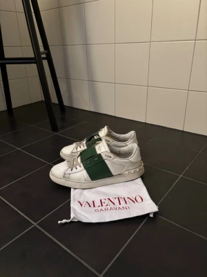 Valentino Garavani sneakers vit/grön - Säljer ett par snygga Valentino Garavani sneakers i vitt med en bred grön rem över mitten. Skorna har rund tå, snörning och coola nitar på sulan. Materialet är skinn Perfekta för dig som gillar exklusiva märken och streetstyle.