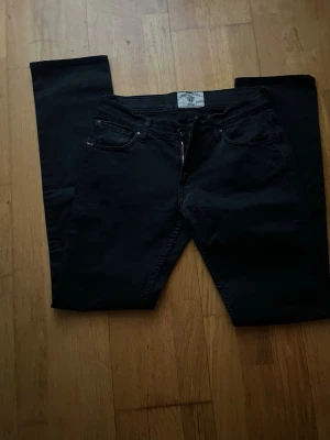 Svarta  jeans från Tiger of Sweden - Säljer ett par svarta Replay Anbass jeans med klassisk femficksdesign och smal passform. Jeansen har dragkedja och knapp framtill, samt Replay-logga på bakfickan. Materialet är mjukt och stretchigt jeans, perfekt för en cool och avslappnad stil.