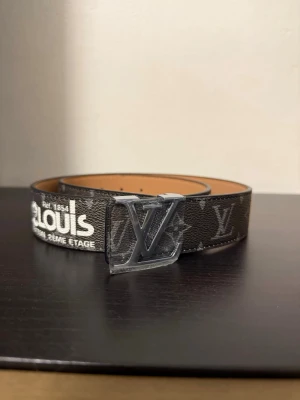 Bälte - Säljer ett snyggt bälte med klassiskt monogrammönster i mörkbrunt och grått. Bältet har en stor spänne i silver och detaljer med vit och gul text. Insidan är beige och materialet är skinn. Perfekt för att lyfta din outfit.