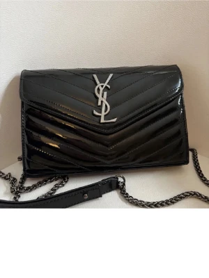 Svart YSL axelväska i skinn - Säljer en snygg svart axelväska från Saint Laurent med kedjerem och silverfärgat YSL-logo på framsidan. Väskan är tillverkad i glansigt skinn med quiltad design och har magnetlås samt flera fack inuti. Perfekt för dig som gillar lyxiga detaljer och stilren look.