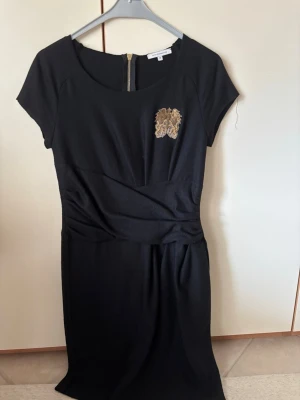 Svart kortklänning från Faith Connexion - Svart kortklänning från Faith Connexion med guldfärgad broderad detalj på bröstet och synlig dragkedja i ryggen. Klänningen har korta ärmar och en snygg drapering i midjan som ger en cool siluett. Perfekt för dig som gillar stilrena och unika plagg.