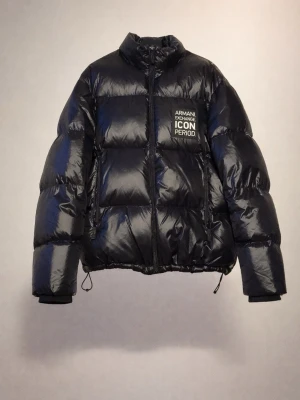 Svart pufferjacka från Armani Exchange - Svart pufferjacka från Armani Exchange med glansig finish och tryckt logga på bröstet. Jackan har hög krage, dragkedja framtill och två sidofickor med dragkedja. Justerbar nederkant med snörning och boxig passform. Perfekt för kalla dagar. Nypris ca. 3800kr                                                                            Modellen är 181cm.