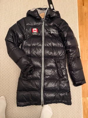 Marinblå lång dunjacka Sail Racing XS - Säljer en mörkt blå lång dunjacka från Sail Racing i storlek XS. Jackan har huva, vit dragkedja, broderad logga och patch med kanadensisk flagga. Den är quiltad och har två sidofickor. Perfekt för kalla dagar när du vill vara både varm och snygg.