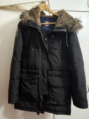 Svart parkajacka med fuskpäls från Bondelid - Parkajacka i svart från Bondelid, modell Cheyenne. Jackan har en stor huva med fluffig fuskpäls, flera fickor med lock och dragkedja framtill. Materialet är polyamid och polyester, perfekt för kalla dagar. Snygg och praktisk design med detaljer som snörning och knappar.