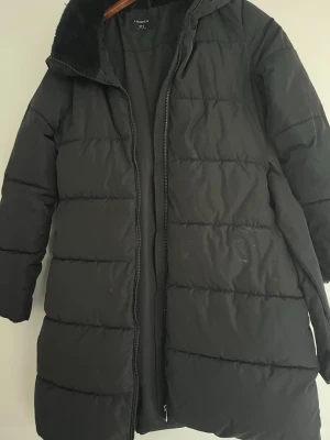 Svart lång pufferjacka från Lindex - Svart pufferjacka från Lindex i storlek S. Jackan är lång, har huva och dragkedja framtill. Perfekt för kalla dagar när du vill vara både varm och snygg.Den har knappt använts, bara några få gånger, och ser ut som ny.