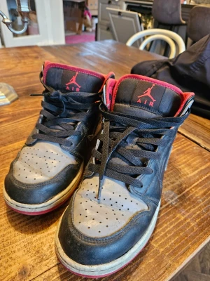 Nike Air Jordan 1 Mid Svart/Röd/Grå - Säljer ett par Nike Air Jordan 1 Mid i svart, grå och röd. Skorna har klassisk snörning, röd insida och yttersula, samt ikoniska Air Jordan-loggan på sidan. Ovandelen är i skinn och syntet, med perforerad tå och vit mellansula. Perfekta för dig som gillar streetwear.