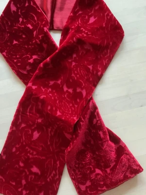 Röd sammetsscarf från NY&CO - Snygg röd scarf i sammet från NY&CO med paisleyinspirerat mönster. Materialet är en mix av rayon och silke med ett lent foder i polyester. Perfekt accessoar för att lyfta din outfit med en lyxig touch och djup färg.