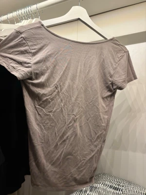 Beige t-shirt med öppen rygg - En enkel beige t-shirt med öppen rygg från lager 157 i storlek S. T-shirten har rundad, djup urringning och korta ärmar. Tillverkad i mjukt bomullsmaterial och har en klassisk, avslappnad passform. Perfekt till jeans eller kjol för en clean look. (OBS har en fläk som inte går bort: se på bilderna)