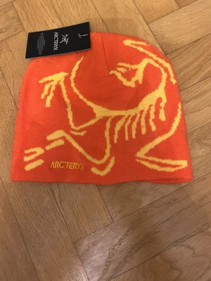 Orange Arc’teryx mössa - Bilderna visar en orange mössa från Arc'teryx med gul logotyp och mönster, inte en väst. Kan ej ge beskrivning eller taxonomier för väst.