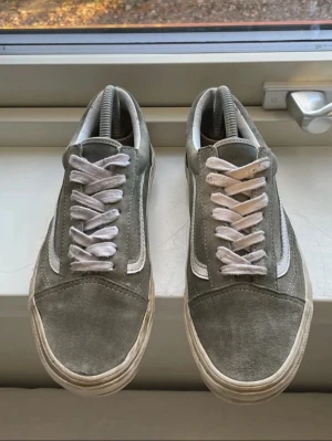 Grå Vans Old Skool sneakers - Säljer ett par grå Vans/gröna Old Skool sneakers med vit sula och vit sidostripes. Skorna har snörning och är tillverkade i mocka med klassisk låg profil och rund tå. Perfekt för dig som gillar streetwear och vill ha en stilren look.