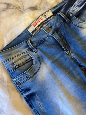 Blå Diesel jeans med slitningar - Säljer ett par blå jeans från Diesel med snygga slitningar och tvättade detaljer. Klassisk femficksmodell med raka ben och kontrastsömmar. Bakfickorna har broderade detaljer och logotyp. Perfekt för dig som gillar en cool och avslappnad stil.