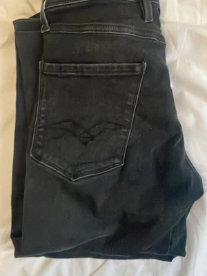 Svarta Replay jeans slim fit - Säljer ett par svarta jeans från Replay med klassisk femficksdesign och snygga detaljer på bakfickorna. Jeansen har slim passform och är tillverkade i mjukt jeansmaterial med stretch. Perfekt för dig som gillar stilrena och bekväma byxor.