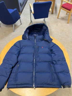 Mörkblå pufferjacka från Polo Ralph Lauren - Säljer en mörkblå pufferjacka från Polo Ralph Lauren med huva och tvåvägsdragkedja. Jackan har en röd Polo-logga på bröstet, praktiska fickor med dragkedja och quiltad design. Perfekt för kalla dagar och riktigt snygg till streetwear.