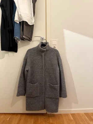 Grå teddyjacka med hög krage - Mysig grå teddyjacka med hög krage och två stora fickor framtill. Jackan har långärmade ärmar och är tillverkad i ett fluffigt, ulligt material som ger en cool och avslappnad vibe. Perfekt för kyliga dagar när du vill vara både varm och snygg.