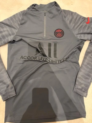Grå Nike PSG träningströja S - Säljer en grå träningströja från Nike med PSG-logga och röd swoosh på ärmen. Tröjan har halv dragkedja, svarta tryck på bröstet och ryggen samt mönstrade ärmar. Materialet är polyester och designen är slim fit, perfekt för fotbollsträning.
