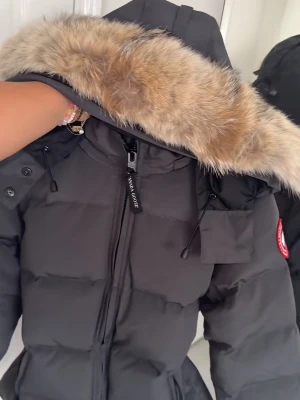 Svart Canada Goose pufferjacka med päls - Säljer en svart pufferjacka från Canada Goose med fluffig päls på huvan. Jackan har dragkedja framtill, praktiska fickor och justerbara snören vid huvan. Perfekt för kalla dagar och har en snygg, klassisk look med märkets ikoniska patch på ärmen. Prislappen är på jackan.