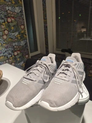 Adidas Asweerun Löparskor Grå Strl 44 - Säljer ett par snygga grå adidas sneakers med mesh-ovandel och vita sulor. Skorna har blå detaljer vid hälen och klassiska adidas-ränder i silver på sidorna. Perfekt för dig som gillar sportig stil och vill ha bekväma skor med snörning.