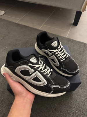 Svarta CD B30 sneakers - Säljer ett par svarta CD B30 sneakers med vita och grå detaljer. Skorna har en chunky vit och svart sula, snörning och stora CD-loggor på sidan och hälen. Ovandelen är i mesh och syntet med coola kontrastpaneler. Perfekt för dig som gillar lyxiga och trendiga sneakers.
