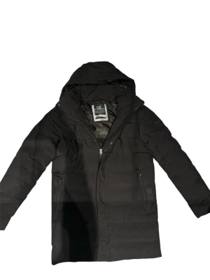 Svart lång dunjacka från Cutter and Buck - Svart dunjacka från Cutter and buck med huva och dragkedja framtill. Jackan är lång, har två sidofickor med dragkedja och en snygg logotyp på ärmen. Perfekt för kalla dagar och har en stilren look som funkar till många outfits. Nypris ca 2200kr