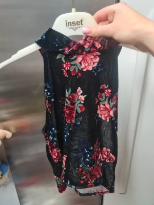 Blommig ärmlös topp från Divided - Säljer en svart ärmlös topp från Divided med blommigt mönster i rött, rosa och blått. Toppen har en hög krage och är gjord i ett mjukt, glansigt polyestermaterial. Perfekt för dig som gillar färgstarka detaljer och blommiga prints.