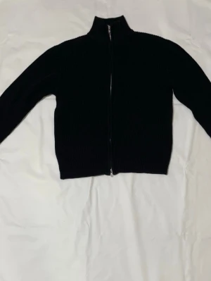 Svart stickad kofta från 157 - Svart ribbstickad kofta från 157 med hel dragkedja framtill och hög krage. Tröjan är långärmad och har en clean, enkel look som passar perfekt till lager-på-lager. Materialet är stickat och känns både mjukt och rejält.