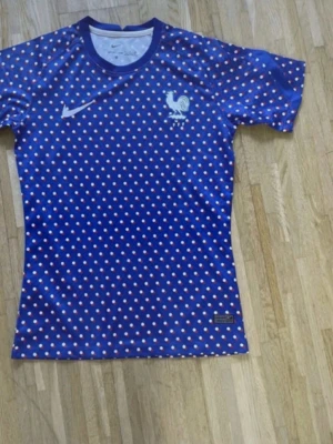 Blå prickig Nike fotbollströja - Säljer en blå fotbollströja från Nike med vitt och rött prickigt mönster över hela tröjan. Tröjan har korta ärmar och rund hals, samt en vit Nike-logga och en tupp på bröstet. Perfekt för dig som gillar sportig stil och vill sticka ut på planen.