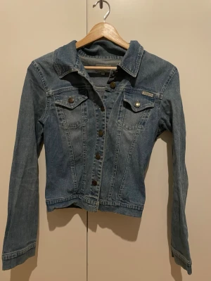 Blå jeansjacka från Tessie - Säljer en klassisk blå jeansjacka från Tessie med snygga bronsfärgade knappar och två bröstfickor med lock. Jackan har en rak passform och långa ärmar, perfekt att slänga över en hoodie eller t-shirt. Enkel och tidlös design som alltid funkar. 