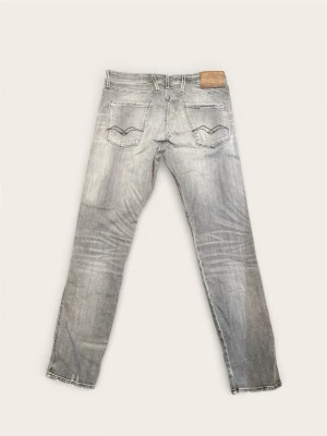 Replay Anbass W34 L32 - Säljer ett par Replay Anbass slim fit jeans i grå tvätt med slitningar och distressed detaljer. Jeansen är tillverkade i ekologisk bomull och har klassiska fem fickor, snygga sömmar på bakfickorna och knappgylf. Perfekta för en avslappnad streetstyle.