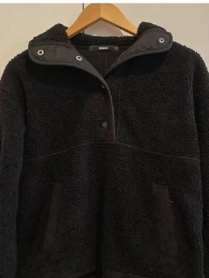 Svart teddyjacka från BikBok XS - Mysig svart teddyjacka från BikBok i storlek XS. Jackan har hög krage med tryckknappar, två fickor framtill och en oversized look. Perfekt för kalla dagar och riktigt skön att bära tack vare det fluffiga materialet.