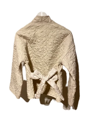 Beige quiltad overshirt med bälte - Quiltad beige overshirt med omlott-design och bälte i midjan. Jackan har lång ärm, två fickor fram och ett blommigt mönster på vissa delar. Perfekt för dig som gillar chill och stilren vibe med extra detaljer. Få 2 i 1 med denna fina jacka! 