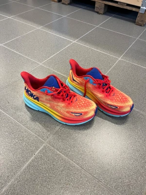 Helt nya  Hoka löpar skor i stl 41  - Säljer ett par Hoka  Clifton 9 sneakers i en riktigt färgstark kombo av rött, gult och blått. Skorna har mesh-ovandel, tjock plattformssula i blått och gult, samt snörning. Perfekta för dig som gillar att sticka ut och vill ha grym komfort till träningen.