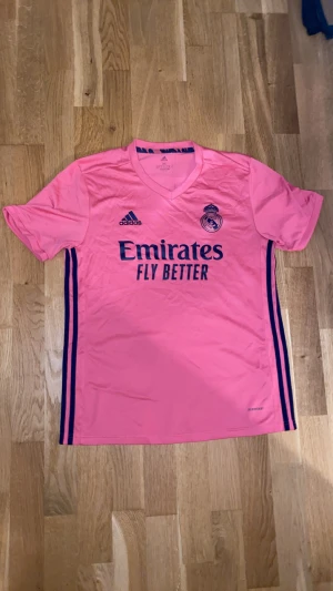 Rosa Real Madrid bortatröja – Adidas - Stilren rosa bortatröja från Real Madrid med mörkblå detaljer. Utrustad med Adidas-logga, Real Madrid-emblem och sponsortrycket Emirates Fly Better på bröstet. Lätt och bekväm tröja som passar både för match, träning och vardagsbruk. Ett modernt och unikt plagg för alla Real Madrid-fans.