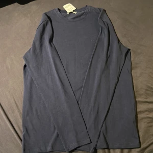 Mörkblå långärmad tröja från ARKET - En stilren mörkblå långärmad tröjai mjuk bomull. Tröjan har en klassisk rund halsringning och enkel design utan tryck eller mönster, perfekt för en clean och avslappnad look. Perfekt för dig som vill vara lite mer grish