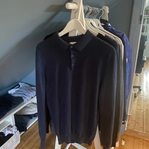 Mörkblå pikétröja i merinoull John Henric - Snygg mörkblå långärmad pikétröja från John Henric i mjuk merinoull. Klassisk krage med tre knappar framtill och ribbade muddar vid ärmslut och nederkant. Perfekt för dig som gillar stilrena och bekväma plagg.