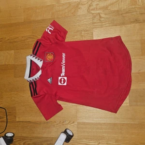 Manchester United röd Adidas fotbollströja XS - Säljer en officiell Manchester United fotbollströja från Adidas i rött med svarta detaljer och vit krage med grafiskt mönster. Tröjan har korta ärmar, broderat klubbmärke och sponsortryck på bröstet. Materialet är lätt och andas, perfekt för match eller träning.