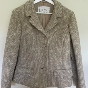 Beige rutig ullkavaj från Wettergrens - Klassisk beige kavaj från Wettergrens i äkta norsk Sandnes tweedull. Snyggt rutigt fiskbensmönster, knappar framtill och två fickor med lock. Perfekt för dig som gillar tidlös stil och vill ha en jacka med schysst passform och cool vintagekänsla.