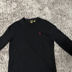 Svart långärmad tröja Polo Ralph Lauren - Svart långärmad tröja från Polo Ralph Lauren i klassisk fit. Tröjan har rund hals och är gjord i mjuk bomull. På bröstet finns den ikoniska röda Polo-loggan broderad. Perfekt för dig som gillar stilrena och tidlösa plagg.