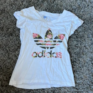 Vit Adidas t-shirt med blommigt tryck - Vit t-shirt från Adidas i 100% bomull med blommigt tryck på loggan och texten. T-shirten har korta ärmar och v-ringad hals. Perfekt för dig som gillar sportiga märken med en twist av färg och mönster.