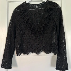 Svart spetsblus med volang från Zara - Supersnygg svart blus från Zara i spets med unika mönster och volangdetaljer framtill. Croppad modell med lång ärm och rund hals. 💓💓