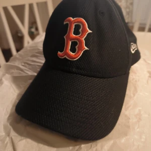 Svart Boston Red Sox keps från New Era - Säljer en svart keps från New Era med Boston Red Sox-logga broderad i rött och vitt framtill. Kepsen är tillverkad i polyester och har klassisk böjd skärm. MLB Genuine Merchandise-märke på insidan. Perfekt för dig som gillar baseball eller vill ha en sportig stil.